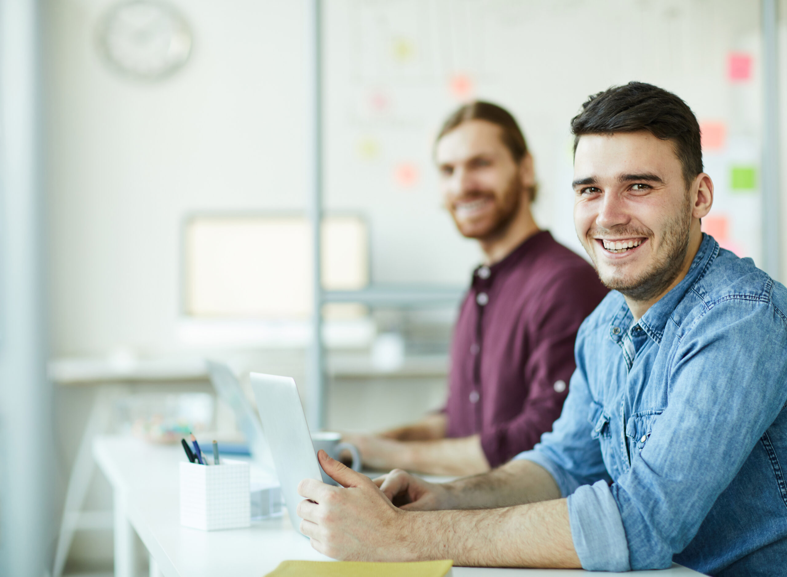 Cheerful young manager with toothy smile and his colleague in casualwear looking at you by workplace