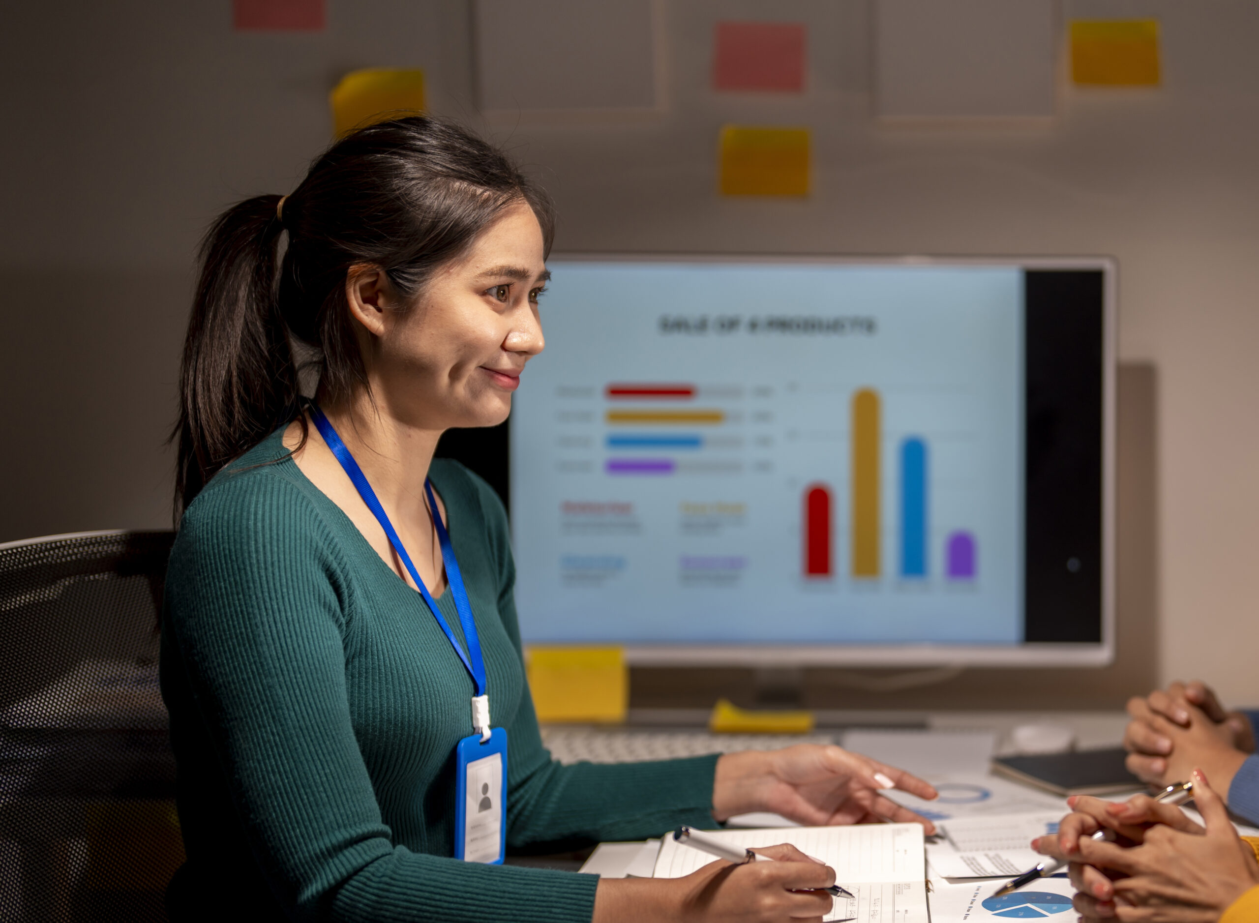 Two women are sitting at a table with a large monitor in front of them. They are discussing something on the screen
